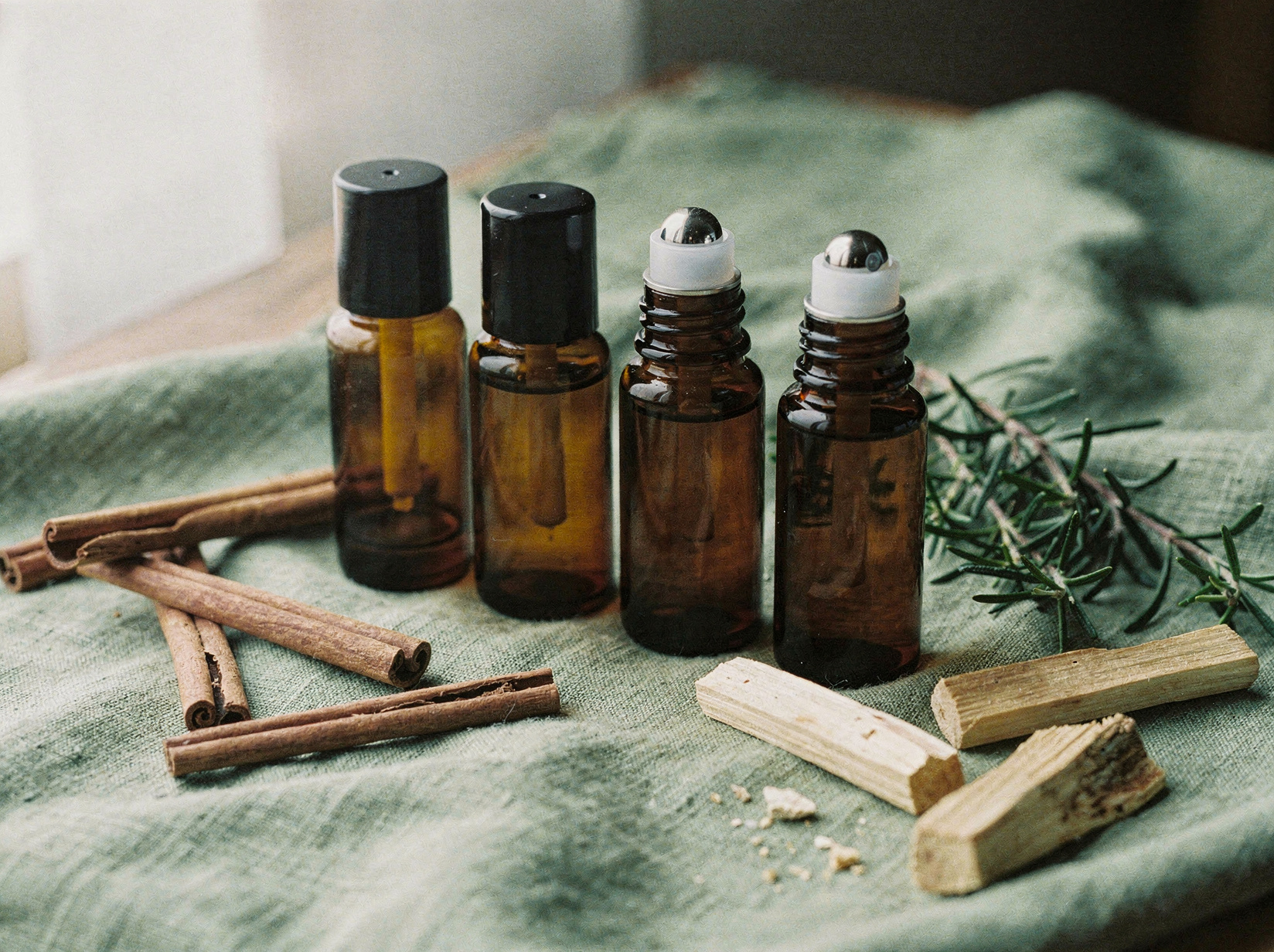 Temporary photo: four amber glass 10ml roll-on bottles with black tops on sage linen with cinnamon, rosemary and palo santo