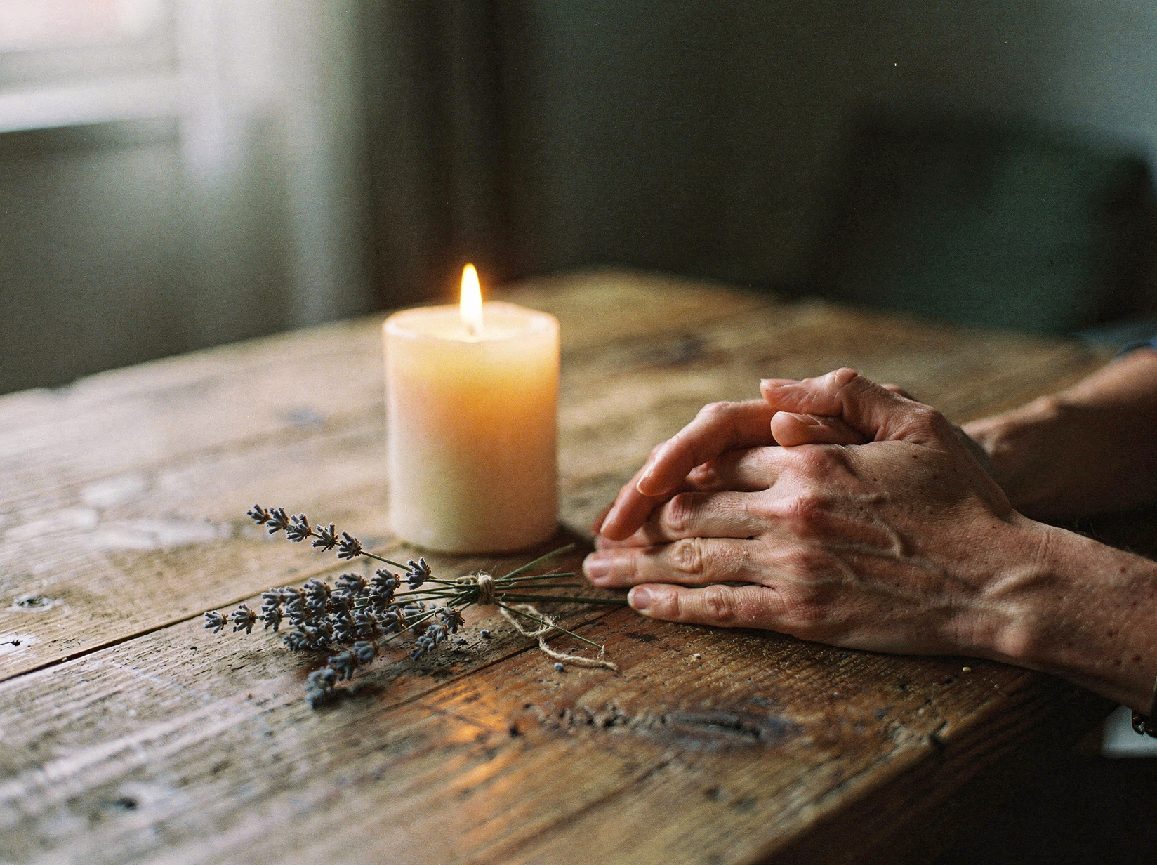 Candle glowing beside lavender and folded hands on reclaimed wood